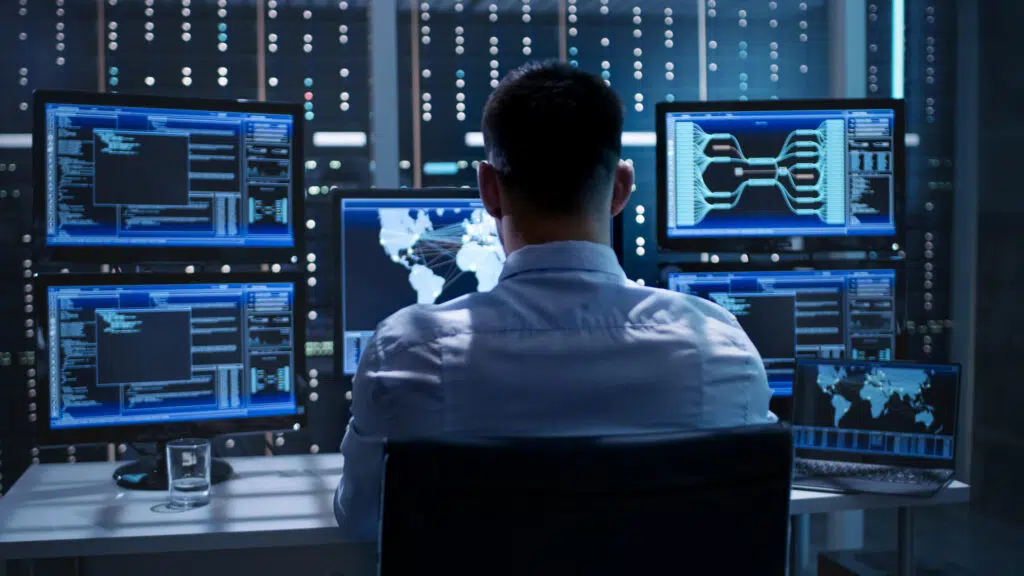 A man sitting in a control room in front of computer monitors with maps and code on the screens