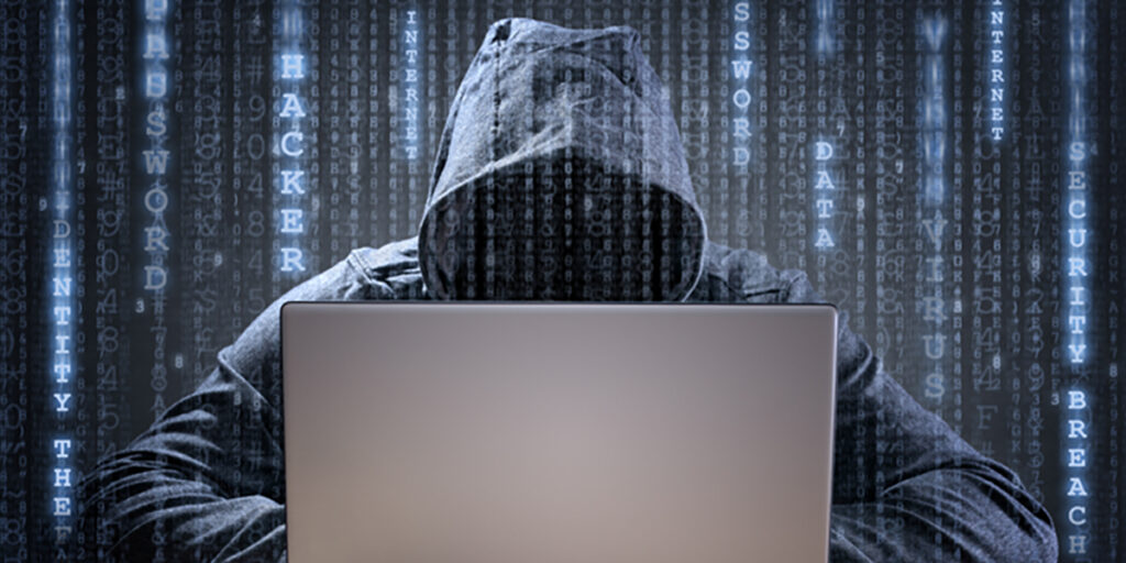 A hooded man sitting with a laptop with code graphics overlaying the image