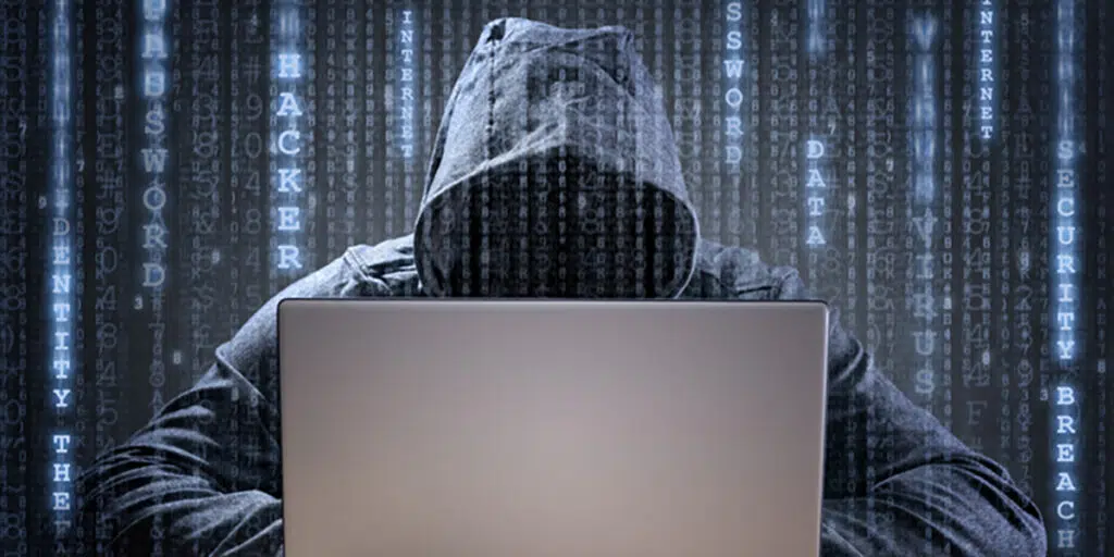 A hooded man sitting with a laptop with code graphics overlaying the image