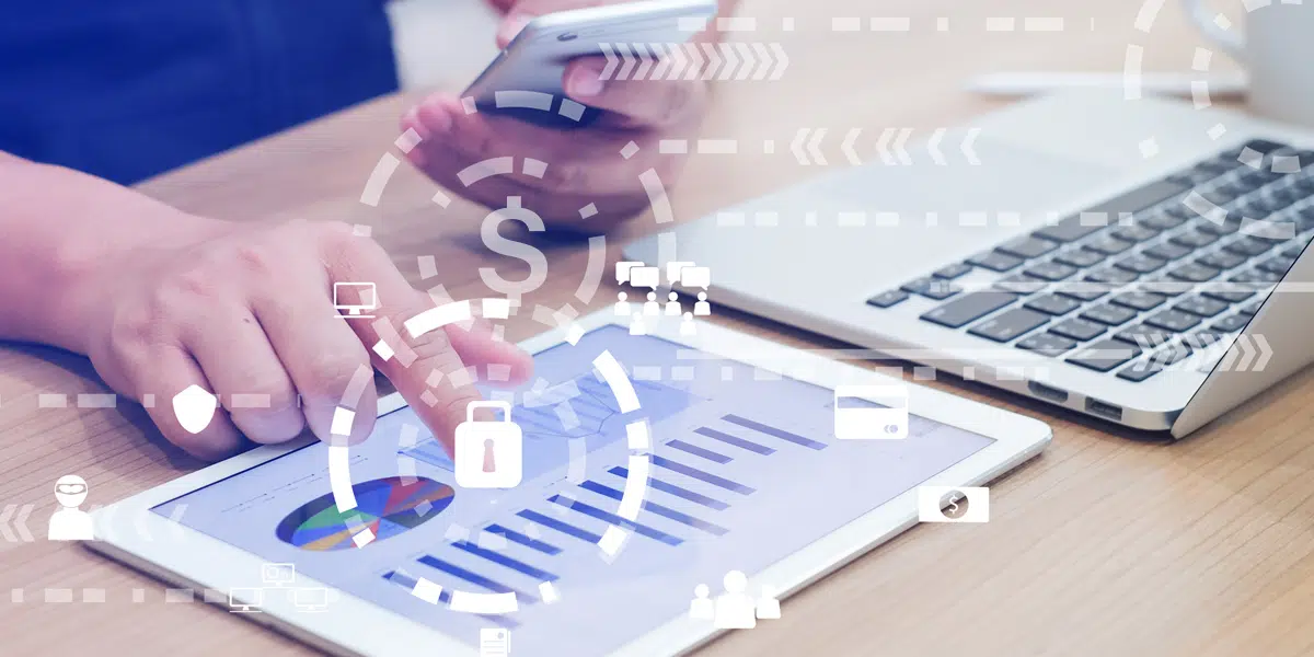 A person holding a cell phone, looking at data on a tablet, with a laptop on a table.